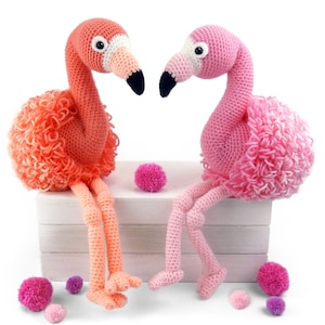May include: Two crocheted flamingo toys, one pink and one orange, sitting on a white shelf with pink pom-poms scattered around them.