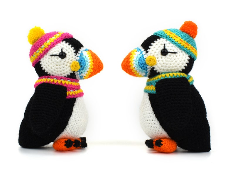May include: Two crocheted puffins wearing colorful hats and scarves. The puffins are facing each other and have black and white bodies with orange beaks.