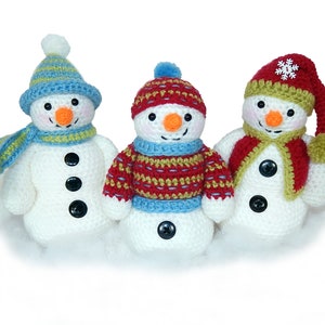 May include: Three crocheted snowmen wearing different hats and scarves. The snowman on the left is wearing a blue and green striped hat and a green and blue scarf. The snowman in the middle is wearing a red and green striped sweater and a blue and green striped scarf. The snowman on the right is wearing a red hat with a white snowflake and a green and red scarf.