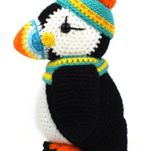 May include: A crocheted stuffed puffin wearing a blue and yellow hat with an orange pom-pom. The puffin has a white body, black wings, and an orange beak.