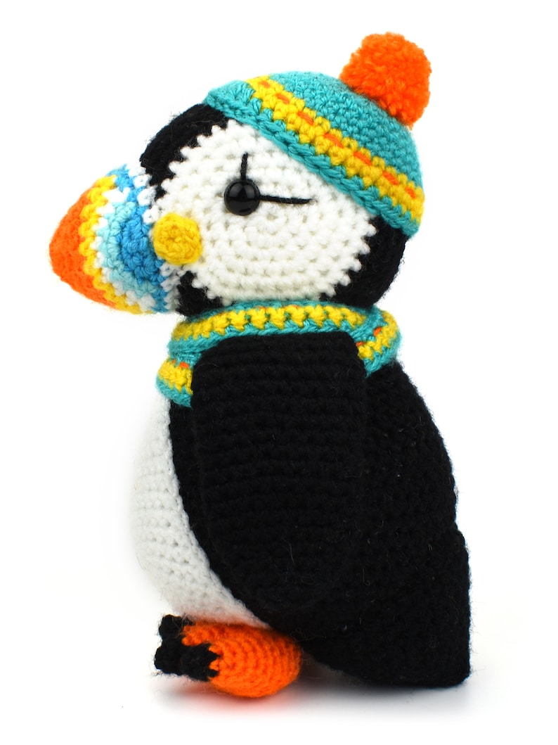 May include: A crocheted stuffed puffin wearing a blue and yellow hat with an orange pom-pom. The puffin has a white body, black wings, and an orange beak.