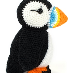 May include: A crocheted stuffed puffin toy with black and white body, orange feet, and a colorful beak with blue, yellow, and orange stripes.