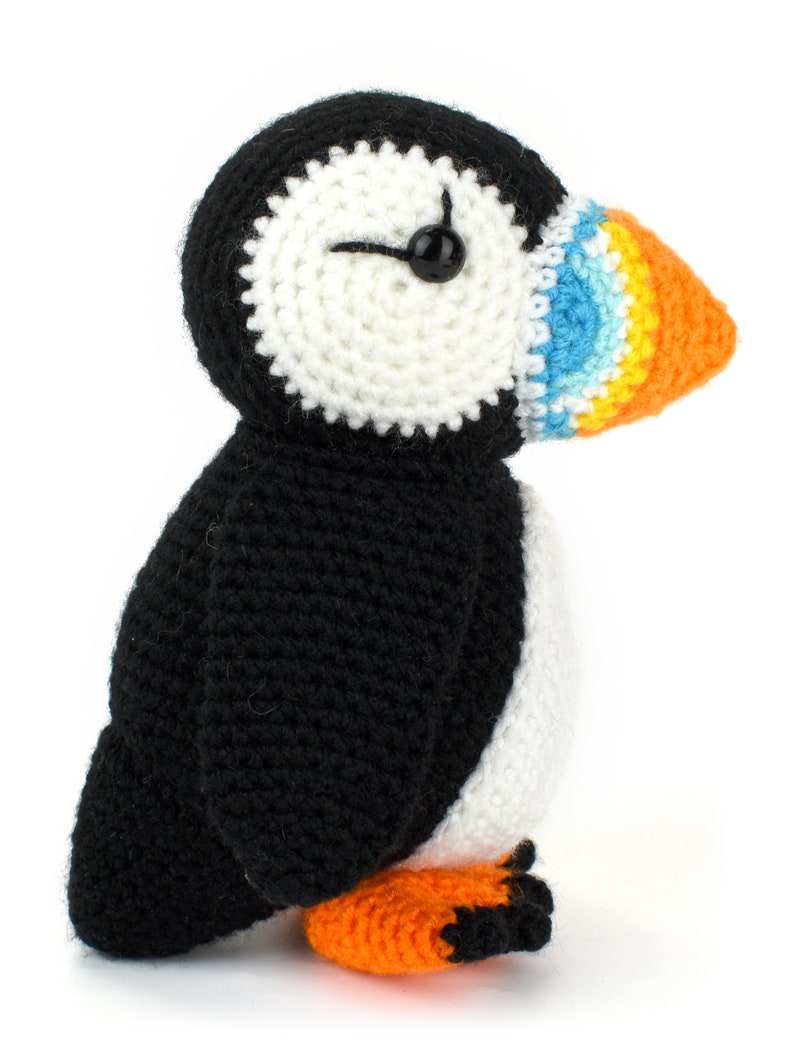May include: A crocheted stuffed puffin toy with black and white body, orange feet, and a colorful beak with blue, yellow, and orange stripes.