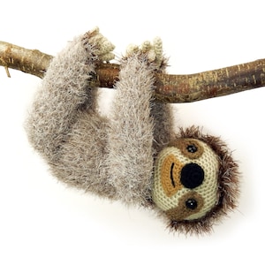 May include: A crocheted sloth hanging upside down from a branch. The sloth has a brown and beige fur, black eyes, and a brown nose.