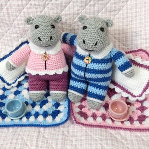 May include: Two crocheted hippopotamus toys wearing pajamas, one in pink and white stripes and the other in blue and white stripes, are lying on a bed with crocheted blankets and pillows. There are two small teacups on the bed.