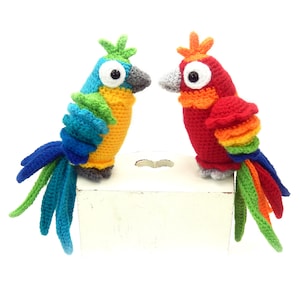 May include: Two crocheted parrots, one blue and yellow, the other red and orange, with colorful tails, sitting on a white surface.