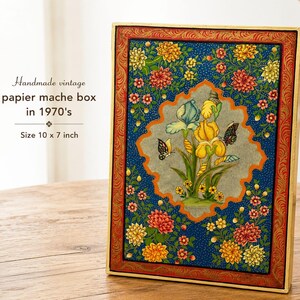 May include: A rectangular papier-mache box measuring 10 by 7 inches. The surface features a dark blue background with colorful floral patterns, a central scalloped panel with yellow and blue irises, butterflies, and a red decorative border.