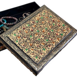 May include: A rectangular black jewellery box with a hinged lid featuring an intricate gold-toned tree design with small green and red accents. The open box reveals a dark interior containing several beaded bracelets and metallic rings.