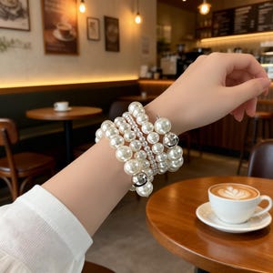 May include: A wrist is adorned with multiple pearl bracelets of different sizes and designs. The bracelets incorporate large, medium, and small faux pearls, alongside silver-coloured metal details. The jewellery is set against a blurred cafe backdrop.