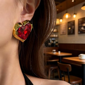 May include: A gold-toned heart-shaped earring with a large, faceted red gemstone center. The earring is worn on the ear, showcasing the bold design and vibrant color. The earring's gold frame adds a touch of elegance.