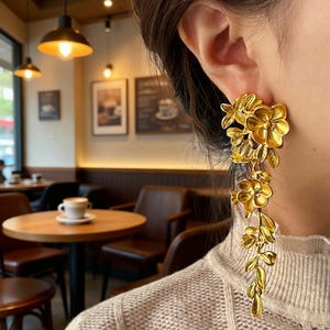 May include: Gold-coloured floral earrings. The earrings feature a cluster of flowers and leaves at the top, with a cascading design of additional flowers and foliage. The earrings are worn on the ear, showcasing their intricate details and design.