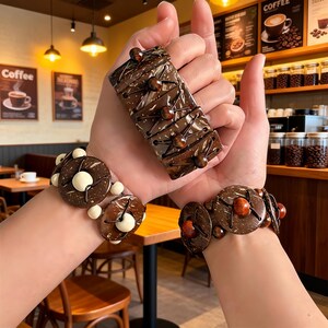 Può includere: Una collezione di gioielli in legno marrone, tra cui un braccialetto largo e due braccialetti di perline. Il braccialetto largo è rettangolare con dettagli marrone scuro e piccole perline marroni. I braccialetti di perline presentano perline rotonde in legno e dettagli bianchi.