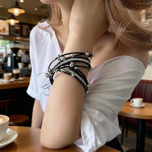 Può includere: Bracciale avvolgente nero con più fili. Il bracciale presenta perline color argento e un filo con piccoli strass trasparenti. Il bracciale è indossato al polso. Lo sfondo è un ambiente da caffè.