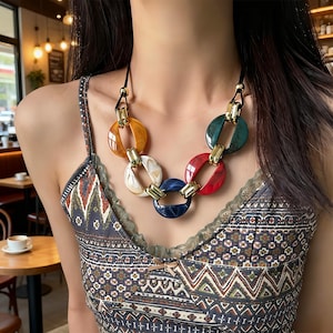 Könnte beinhalten: Eine auffällige Halskette mit großen, kreisförmigen Gliedern in den Farben Bernstein, Weiß, Marineblau, Rot und Grün. Die Glieder sind durch goldfarbene Metallakzente verbunden und an einem schwarzen Band befestigt. Ein auffälliges Modeaccessoire.