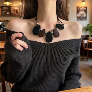 May include: A necklace featuring black and brown teardrop-shaped pendants with silver beads. The necklace is made of polished stones and is strung on a black cord. The necklace is worn over a dark grey off-the-shoulder jumper.