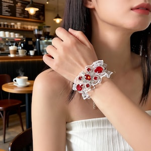 May include: A white lace bracelet adorned with large, vibrant red gemstones and smaller clear crystals. The bracelet features a delicate, ruffled design and is worn on a wrist. The background shows a cafe setting.