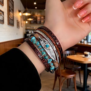May include: A multi-layered bracelet set featuring brown leather, silver, and turquoise accents. The bracelets include a feather charm, braided details, and turquoise stone beads. The set is designed to be worn together for a layered look.