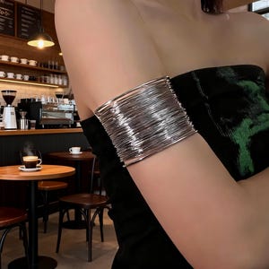 Puede incluir: Un brazalete plateado, compuesto por numerosas bandas metálicas paralelas, se lleva en la parte superior del brazo. El brazalete es de un plateado brillante y reflectante, y las bandas crean una apariencia texturizada y en capas. El fondo muestra un café.