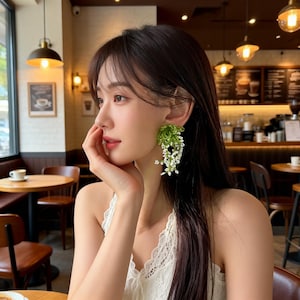 May include: A woman wearing a white lace top and large, unique earrings. The earrings feature a cluster of green foliage with cascading white flowers. The background shows a cafe setting with round tables and hanging lights.