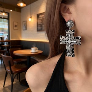 May include: Large cross-shaped earrings with a cowhide pattern and silver-colored metal accents. The earrings have a decorative floral design at the top and center. The earrings are a bold statement piece.