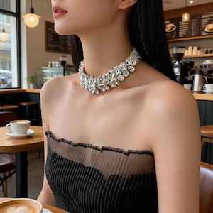 Pode incluir: Um colar marcante prateado brilhante com detalhes de cristal transparente. O colar apresenta um design em forma de folha com pedras grandes e facetadas. A modelo usa um top preto sem alças em um café.