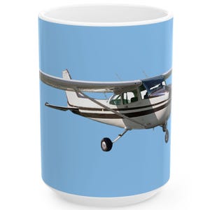 May include: A white ceramic mug featuring a light blue background and a graphic of a small, single-engine propeller airplane. The aircraft is white with dark brown horizontal stripes along the fuselage and has a high-wing configuration.