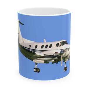 May include: A white ceramic mug featuring a graphic of a twin-engine turboprop airplane in flight against a solid light blue background. The airplane is depicted from a side angle with its landing gear extended and dark blue stripes along the fuselage.