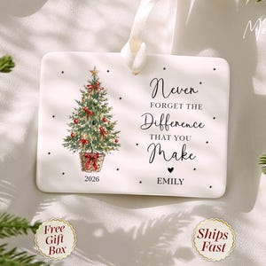 May include: A rectangular white ceramic ornament with a cream ribbon hanger. It features a watercolor Christmas tree in a basket, the text "Never FORGET THE Difference THAT YOU Make", the year "2026", and the name "EMILY" in black.