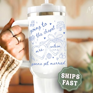 May include: White travel mug with a handle, featuring blue handwritten text and illustrations for a wedding. The text includes "going to the chapel," "Mrs. Wilson," and "gonna get married." A wedding-themed gift.