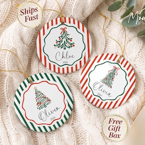 May include: Three round ceramic ornaments with gold hanging strings. Each features a central scalloped frame containing a hand-drawn Christmas tree illustration, a name, and the year 2026. The borders have red or green diagonal stripes.