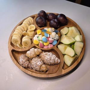 Könnte beinhalten: Ein rundes Serviertablett aus Holz mit fünf unterteilten Fächern, gefüllt mit Bananenscheiben, dunkelvioletten Pflaumen, grünen Apfelspalten, mit Puderzucker bestäubten Hörnchen und einer zentralen Ansammlung bunter Fruchtgummis.