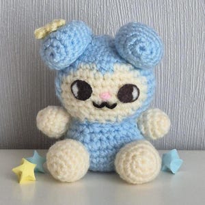 May include: A small, handmade crochet plush toy featuring a light blue body with cream-coloured accents on the face, hands, and feet. The character has large, dark oval eyes, a small pink nose, and a stitched mouth, with a yellow star on one ear.