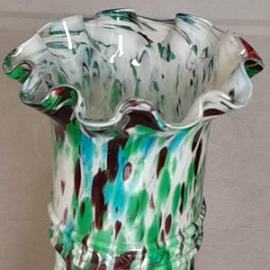 Puede incluir: Un florero de vidrio con base blanca y salpicaduras moteadas en verde, marrón oscuro y azul claro. El florero tiene un borde ondulado y acampanado, y un diseño de cuerpo retorcido en espiral con una banda horizontal en relieve en el cuello.