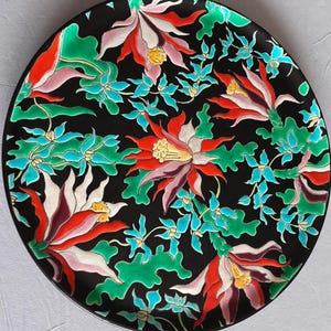 May include: A round black decorative plate featuring a vibrant floral pattern. The design includes large, multi-petaled flowers in shades of red, white, and yellow, interspersed with smaller turquoise blue blossoms and green foliage across the surface.