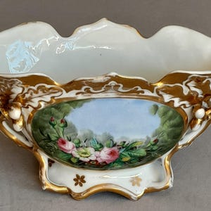 Könnte beinhalten: Eine weiße Porzellanschale mit gewelltem Rand und goldenen Akzenten. In der Mitte befindet sich eine oval handgemalte Szene mit rosa Rosen und grünem Laub vor einem hellblauen Himmel. Kleine weiße beerenartige Details verzieren die Seiten.