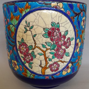 Könnte beinhalten: Ein zylindrischer Keramik-Übertopf mit leuchtend blauem Hintergrund und bunten Blumenmustern. Ein zentrales kreisförmiges Medaillon zeigt eine cremefarbene Glasur mit Rissen und eine detaillierte Abbildung rosafarbener Blüten an braunen Zweigen.