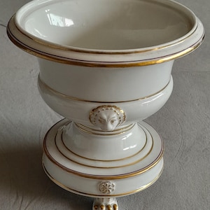 Könnte beinhalten: Eine weiße Keramikurne mit einem breiten, ausgestellten Rand und einem tiefen Becken. Die Außenseite ist mit goldenen Verzierungen, einem skulptierten Löwenkopf-Relief am Hals und einem runden Fuß mit drei goldenen Klauenfüßen versehen.