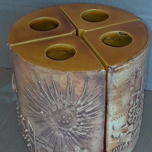 May include: A segmented, ceramic candle holder with four circular openings. The holder is tan and brown with a textured, carved design featuring a sunflower and architectural elements. The candle holder is a decorative home accent.