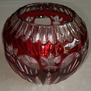 Puede incluir: Un cuenco redondo de cristal tallado en color rojo intenso, con intrincados patrones geométricos y motivos florales cortados a mano. El vidrio transita de un rojo rubí vibrante a secciones transparentes facetadas que captan la luz y reflejan.