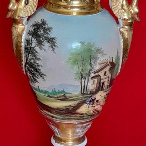 Puede incluir: Un jarrón de porcelana en forma de urna con asas doradas en forma de cisne y un cuello y base pintados en oro. El cuerpo muestra un paisaje pintado a mano con árboles, un edificio y pequeñas figuras en un camino bajo un cielo azul.