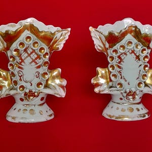 Pode incluir: Um par de vasos de cerâmica branca com detalhes pintados a ouro, bordas recortadas e padrões circulares em relevo. Os vasos possuem topo alargado, corpo central decorado com um design de grade e uma base larga com pontos dourados.