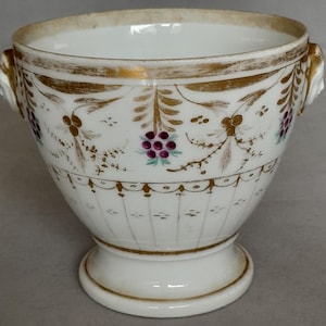 Puede incluir: Un cuenco de porcelana blanca con base de pedestal y dos asas laterales con forma de cabeza de león. El exterior presenta un borde dorado pintado a mano en el borde y la base, con motivos de hojas doradas y racimos de pequeñas bayas moradas.