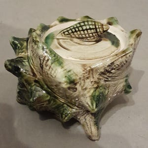 Puede incluir: Un tarro de cerámica con tapa en forma de concha marina espinosa con un acabado brillante en tonos crema, bronceado y verde oscuro. La tapa presenta un pequeño mango texturizado en forma de concha y el cuerpo tiene un exterior irregular.
