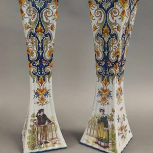 May include: A pair of tall, fluted ceramic vases featuring a white base with intricate blue, orange, and green scrollwork patterns. Each vase displays a detailed illustration of a person in historical attire standing near a wooden fence.