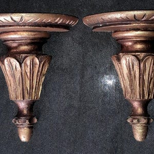 Puede incluir: Dos apliques de pared ornamentados, de estilo antiguo. Cada aplique presenta una repisa circular con detalles tallados, sostenida por un cuerpo acanalado en forma de jarrón. Los apliques están acabados en una combinación de bronce y oro.