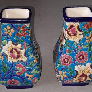 Può includere: Una coppia di vasi in ceramica di forma quadrata con uno sfondo blu vibrante decorato con un colorato motivo floreale di fiori rosa, gialli e verdi. Ogni vaso presenta un bordo blu scuro e un interno bianco, con un collo affusolato.