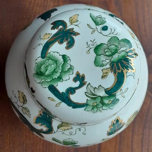 Peut inclure: Un pot en céramique rond avec couvercle, présentant une base de couleur crème ornée de motifs floraux verts complexes, de feuillages en volutes et de contours dorés. Le motif recouvre à la fois le couvercle et le corps du pot.