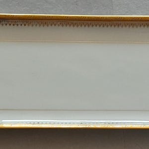 Peut inclure: Un plateau de service rectangulaire en porcelaine blanche doté d'un bord doré poli et de poignées dorées assorties à chaque extrémité. Le plateau présente une surface lisse et brillante avec un fin liseré doré et un motif pointillé délicat.