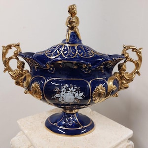 Może przedstawiać: Głęboko niebieska porcelanowa waza z pokrywką zwieńczoną złotą figurką. Naczynie posiada ozdobne złote uchwyty, podstawę w formie cokołu oraz złote wykończenia. Na przedniej części misy namalowany jest biały wzór kwiatowy.