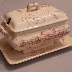 Könnte beinhalten: Eine rechteckige Terrine aus Keramik mit passendem Deckel und Untersetzer. Die cremeweiße, glasierte Oberfläche ist mit einem detaillierten rotbraunen Blumen- und Gräsermuster verziert. Der Deckel verfügt über einen mittigen Griff.